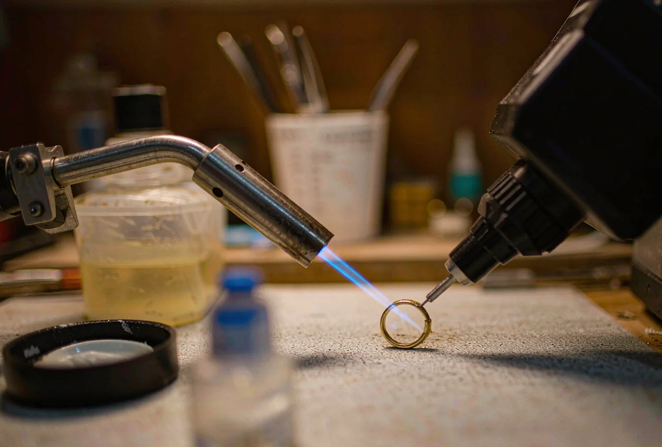 A jeweler's bench with a small gas torch and a laser welding head both aimed at a gold ring
